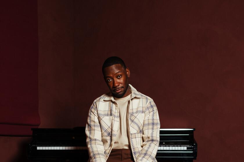 Lamorne Morris sits on a piano bench in a cream flannel and brown pants, with a red backdrop during a 2024 portrait session