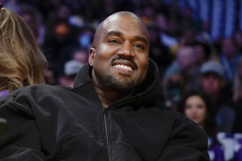 Kanye West leans back as he smiles in a black hoodie while sitting in a crowd